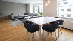 Un salon moderne avec un canapé blanc, une table à manger avec quatre chaises et une grande fenêtre donnant sur des immeubles.