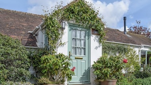 A quaint cottage with a green door, surrounded by lush greenery and potted plants.