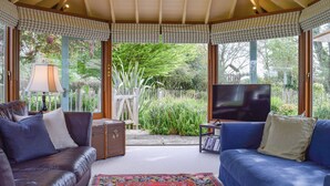 A cozy living room with a large TV, leather sofas, a patterned rug, and a view of a garden through large windows.