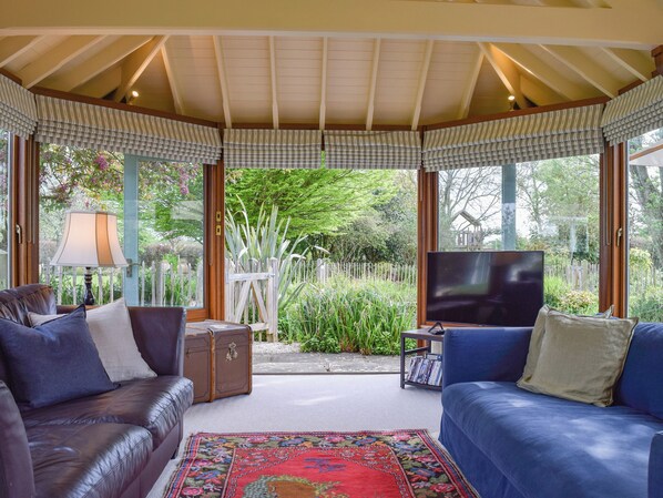 A cozy living room with a large TV, leather sofas, a patterned rug, and a view of a garden through large windows.