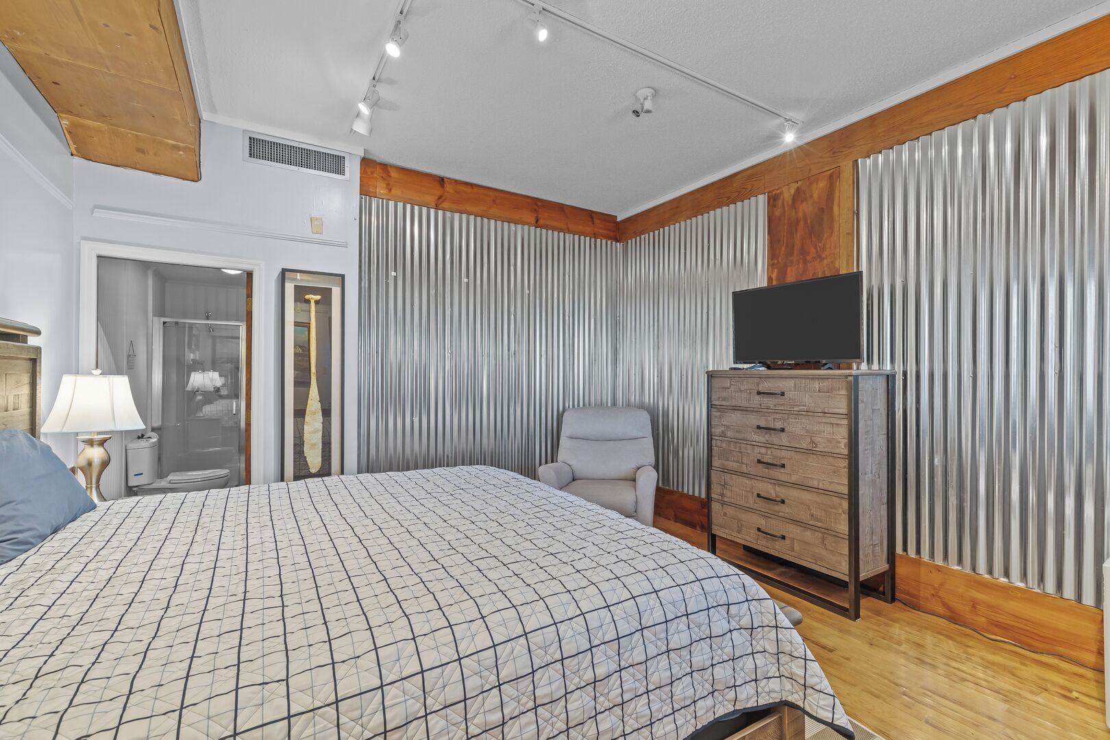 Een slaapkamer met een bed, een dressoir, een televisie, een stoel en een badkamer zichtbaar door een geopende deur.