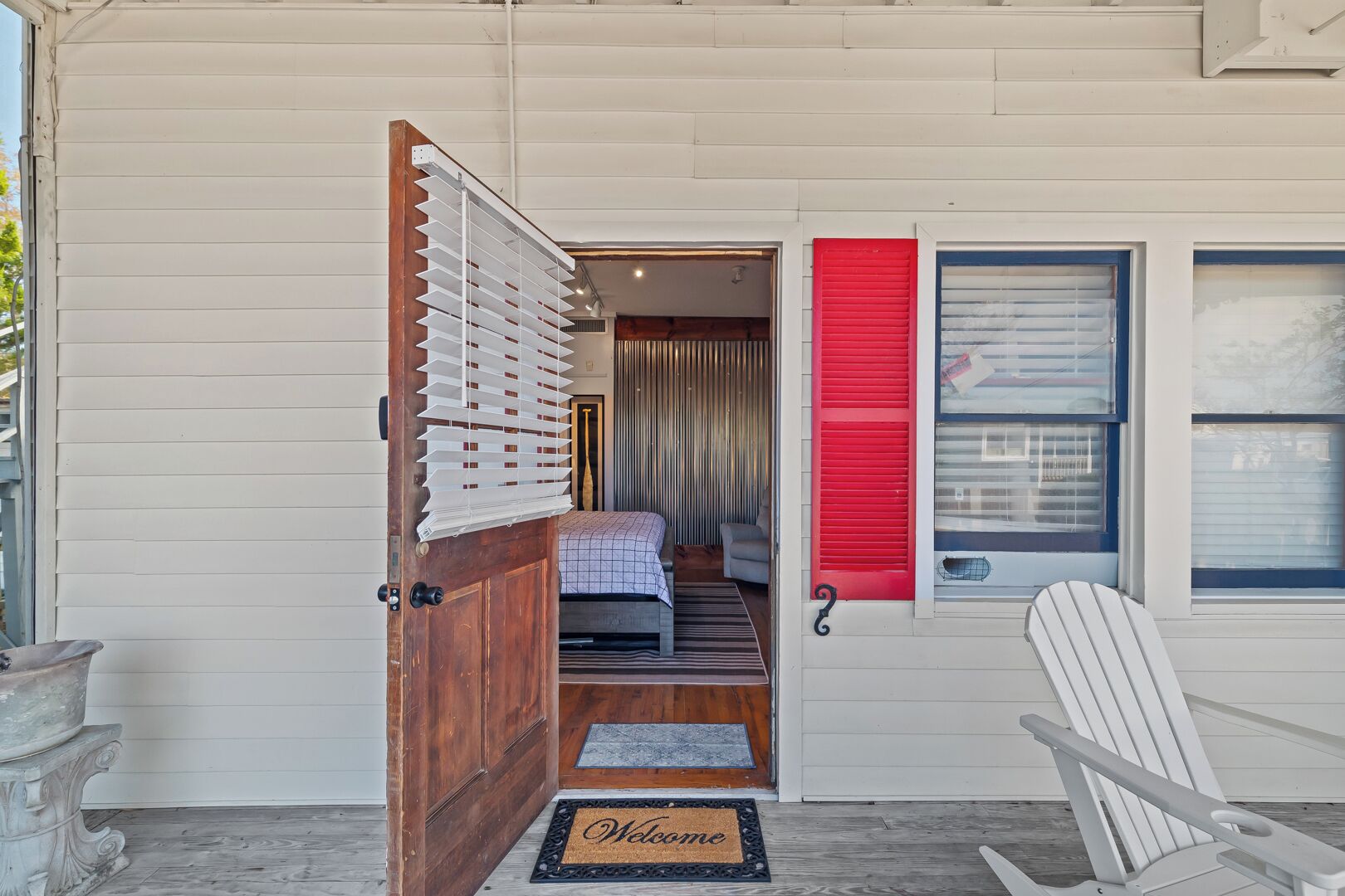Une porte en bois avec un store blanc, un volet rouge et une chaise blanche devant un bâtiment.