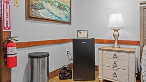 Habitación de hotel con un cuadro de un faro, un extintor, un cartel de Wi-Fi, un refrigerador, una lámpara y un aparador de madera.