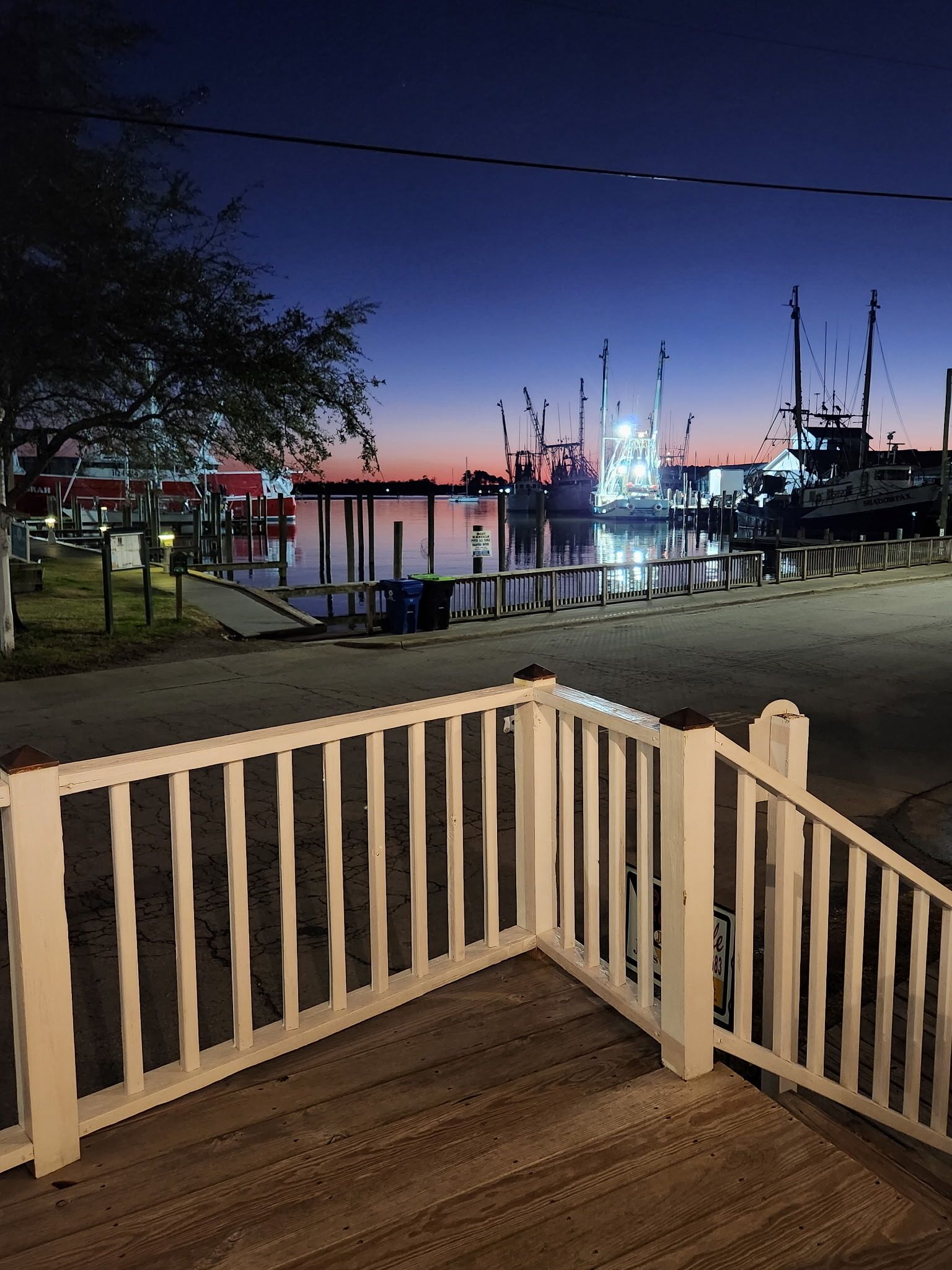Une terrasse en bois avec une rambarde, un quai avec des bateaux, et une étendue d’eau calme au crépuscule.