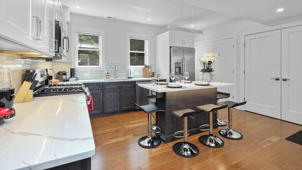 Uma cozinha moderna com armários brancos, uma ilha central com banquetas, eletrodomésticos de aço inoxidável e uma janela que proporciona luz natural.