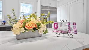Cozinha com armários brancos, geladeira de aço inoxidável e bancada de mármore. Há um buquê de flores sobre a bancada e uma placa de autorização de estacionamento.