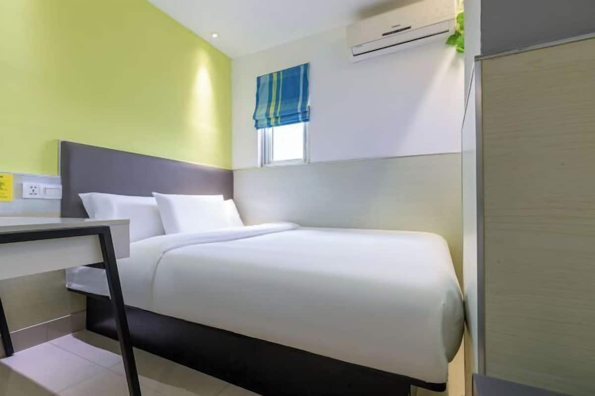 A hotel room with a bed, a bedside table, a window with a blue curtain, and an air conditioning unit.