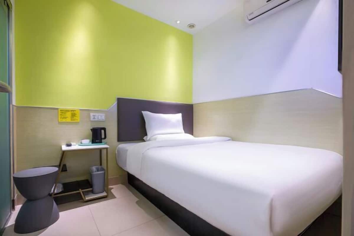 A hotel room with a single bed, a small bedside table with a coffee maker, a trash bin, and a wall-mounted air conditioner.