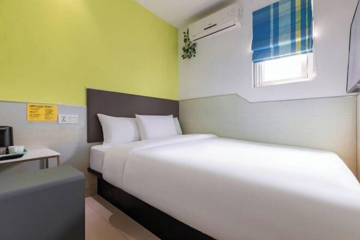 A hotel room with a bed, a small table with a coffee maker, a window with a blue blind, and an air conditioning unit on the wall.