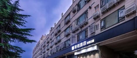 Un hôtel à plusieurs étages, avec une rangée de boutiques au rez-de-chaussée, une entrée bien visible et un espace de stationnement.