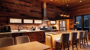 A modern kitchen with wooden cabinetry, a central island with bar stools, and a large window with a view of trees. - Cozy Mountain Getaway with Hot Tub  Sleeps Your Whole Group (Revelstoke)