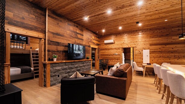 A cozy cabin-style room with a stone fireplace, wooden walls, a TV mounted above it, a sofa, a dining table with chairs, and a bunk bed with a ladder. - Cozy Mountain Getaway with Hot Tub  Sleeps Your Whole Group (Revelstoke)