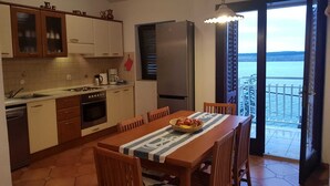 A kitchen with a dining area, a refrigerator, and a view of the sea through an open door.