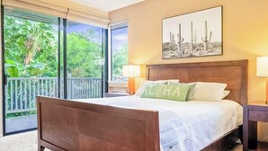 A bedroom with a wooden bed, a view of greenery through large windows, and a cactus-themed artwork on the wall. - Spacious 3BR/2BA Condo in Wailea – Walk to Beach & Shops! (Wailea-Makena)