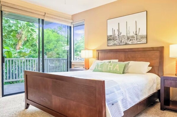 A bedroom with a wooden bed, a view of greenery through large windows, and a cactus-themed artwork on the wall. - Spacious 3BR/2BA Condo in Wailea – Walk to Beach & Shops! (Wailea-Makena)