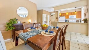A dining area with a wooden table set for four, a mirror on the wall, and a kitchen in the background. - Spacious 3BR/2BA Condo in Wailea – Walk to Beach & Shops! (Wailea-Makena)