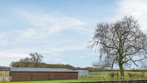 A wooden barn with a sloped roof, a fenced area, and a large leafless tree in the background.