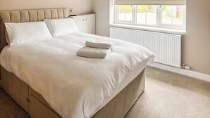A neatly made bed with white linens and two pillows, a beige headboard, a window with blinds, and a radiator.