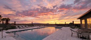 Una piscina con sillas de descanso y sombrillas, rodeada por una valla, con un atardecer al fondo.