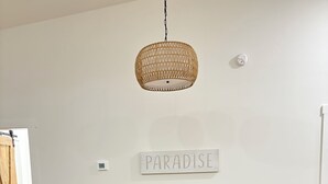 Un comedor con una mesa de madera y sillas de metal negro con asientos trenzados, una lámpara colgante de mimbre y un cartel en la pared que dice "PARADISE".