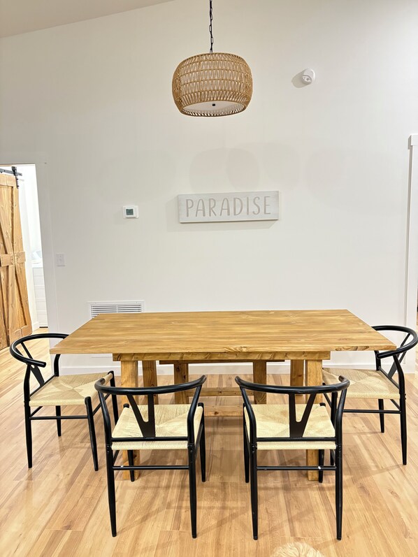 Un comedor con una mesa de madera y sillas de metal negro con asientos trenzados, una lámpara colgante de mimbre y un cartel en la pared que dice "PARADISE".