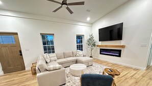 Una sala moderna con un sofá modular, una mesa de centro redonda, un sillón azul, un televisor de pantalla plana sobre la chimenea y piso de madera.