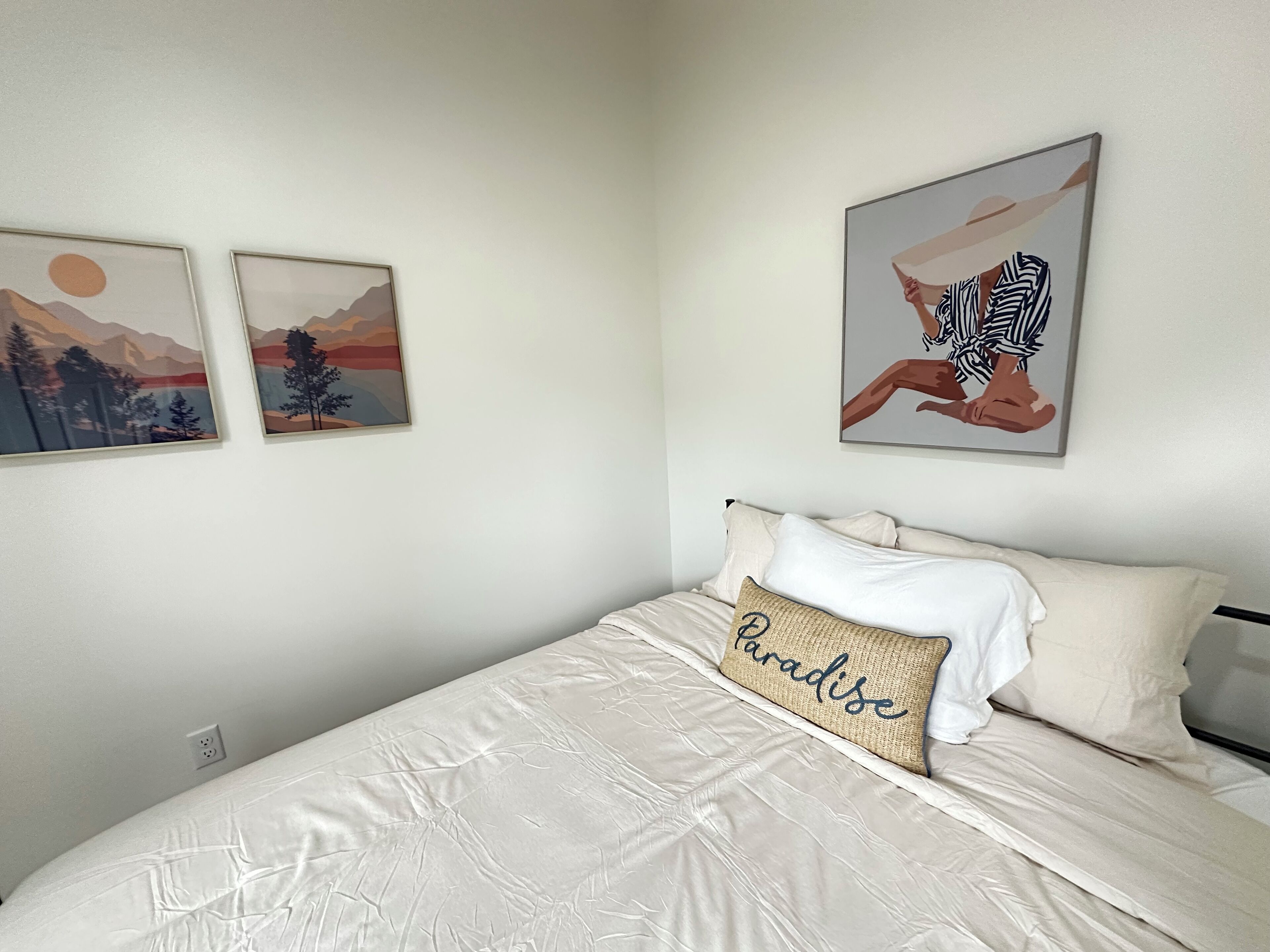A bedroom with a bed, two framed pictures on the wall, and a pillow with the word "Paradise".