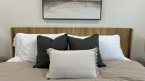 Una cama con cabecera de madera, almohadas blancas y grises oscuras, y una colcha color beige.