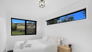 Un dormitorio moderno con una cama grande, una mesita de noche con lámpara, un armario pequeño y dos ventanas con vistas a un área residencial.