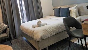 A hotel room with a bed, a desk, a chair, a round wooden table, a chandelier, and a painting on the wall. - RT properties at the Lennox  (Accra)