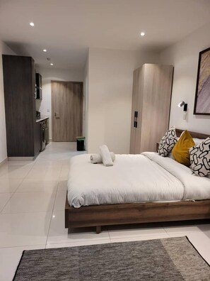A modern bedroom with a large bed, a wooden wardrobe, and a kitchenette in the background. - RT properties at the Lennox  (Accra)