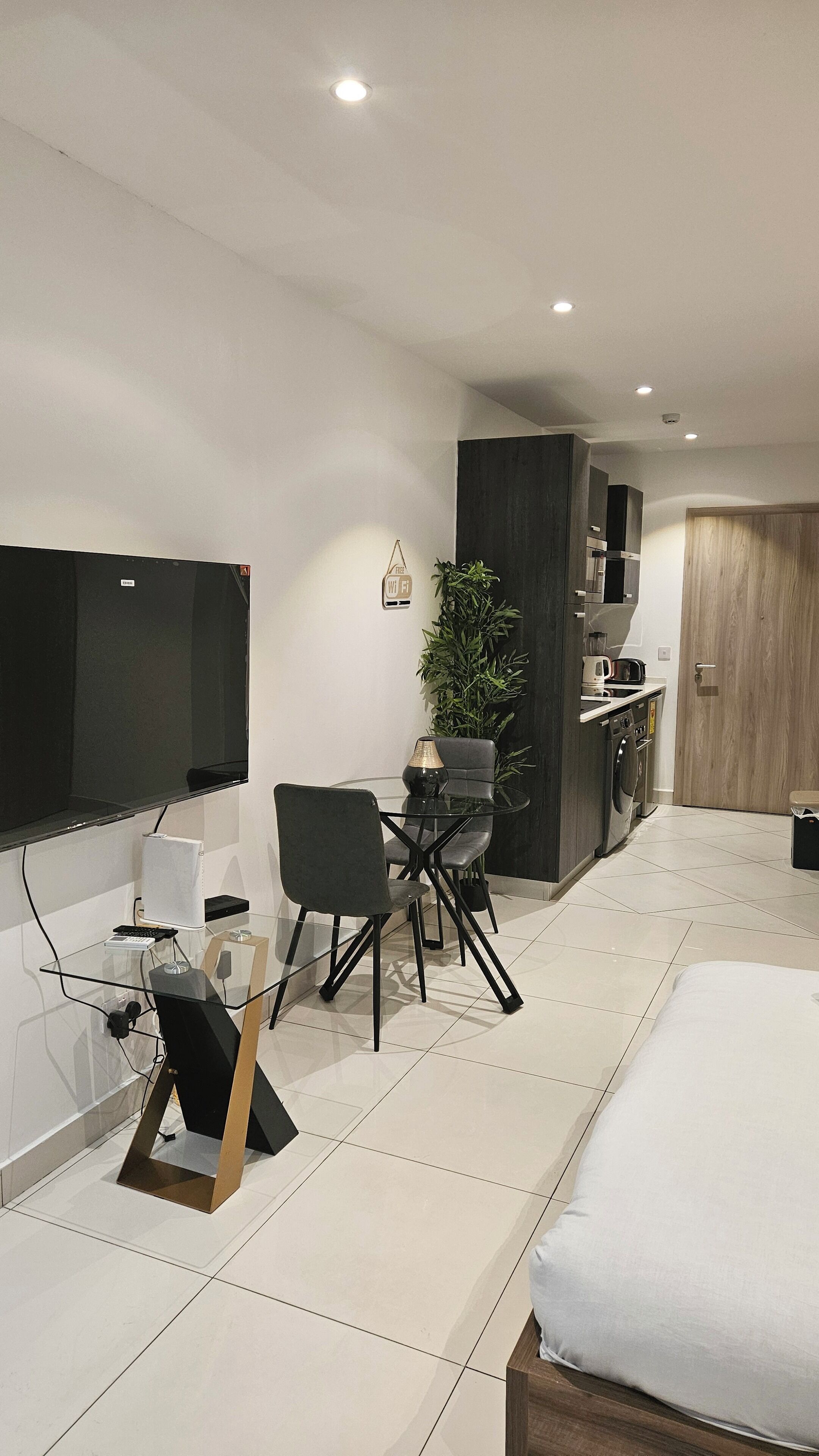 A modern hotel room with a bed, a desk with a chair, a TV, a plant, and a kitchenette.