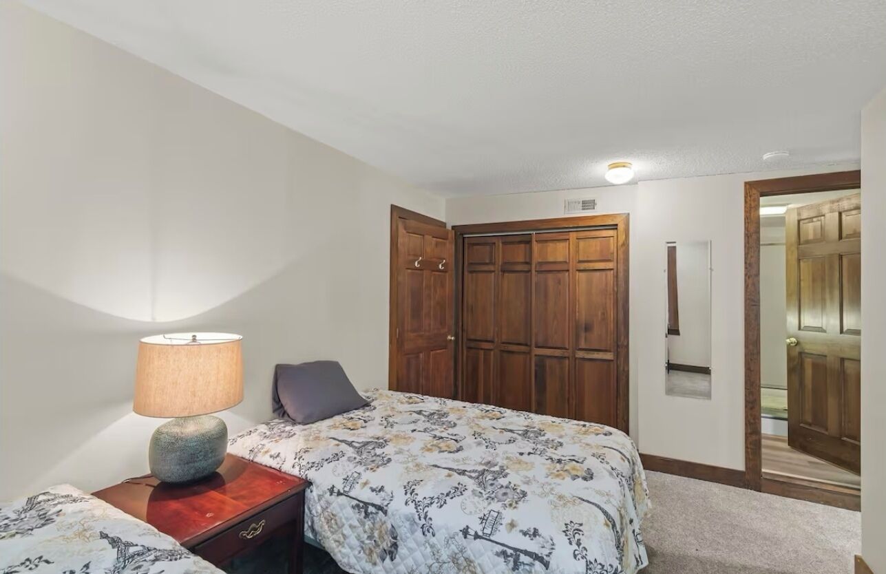 Une chambre à coucher comprenant un lit, une table de chevet avec une lampe, une armoire en bois et une porte donnant sur une autre pièce.