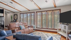 Ein modernes Wohnzimmer mit einem braunen Ledersofa, einem blauen Sofa und einem Couchtisch aus Holz. Ein großes Fenster mit Jalousien, ein Fernseher auf einem weißen Schrank und eine Pflanze auf dem Fensterbrett.