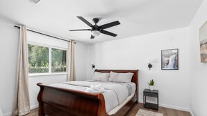 Un dormitorio con cama de madera, ventilador de techo, ventana con cortinas, mesita de noche con una planta y un cuadro en la pared.