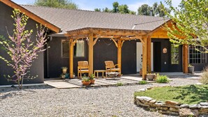Un patio cubierto con sillas y mesas de madera, un pequeño árbol y un borde de piedra.