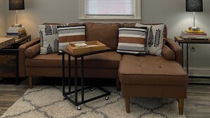 A living room with a brown sectional sofa, a small wooden table, and two side tables with lamps. There are framed pictures on the walls and a window with blinds. - Hip Suite-1st Floor - Arts District/Farmers Mkt/Cafes/Breweries-Near Lake George (Glens Falls)