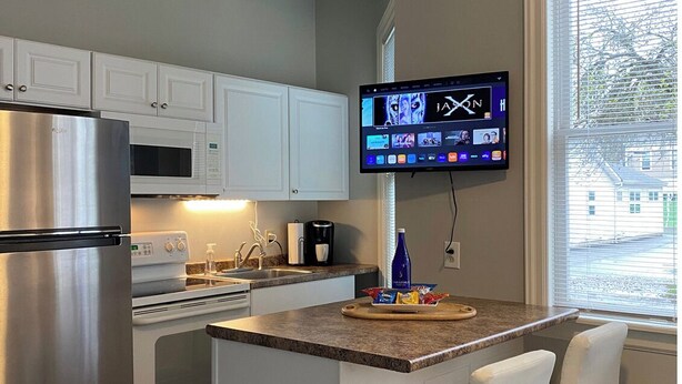 A modern kitchen with white cabinets, stainless steel appliances, a flat-screen TV mounted above the counter, and a window with blinds. - Hip Suite-1st Floor - Arts District/Farmers Mkt/Cafes/Breweries-Near Lake George (Glens Falls)