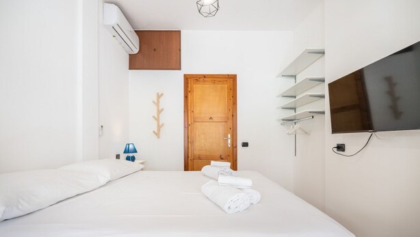 A hotel room with a bed, a TV, a wooden door, and a wall-mounted shelf. - Gigi's House – Comfort & Relax (Villasimius)