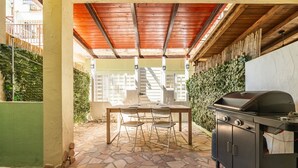An outdoor patio with a stone floor, a wooden table with chairs, a grill, and a covered area with a wooden ceiling. - Gigi's House – Comfort & Relax (Villasimius)