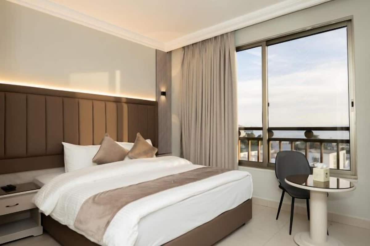 A hotel room with a large bed, a nightstand, a chair, a small table, and a window with a balcony view.