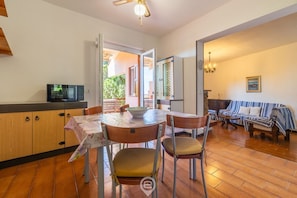 Een eetgedeelte met een tafel, stoelen, een televisie en een keuken met kastjes en een magnetron.