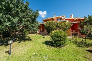 Una casa de dos pisos con fachada roja, una puerta principal visible y un pequeño patio delantero con césped y un árbol.