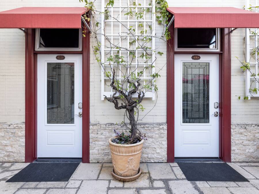 A building with two white doors, each with a red awning and a small window. There is a potted plant in front of the doors.