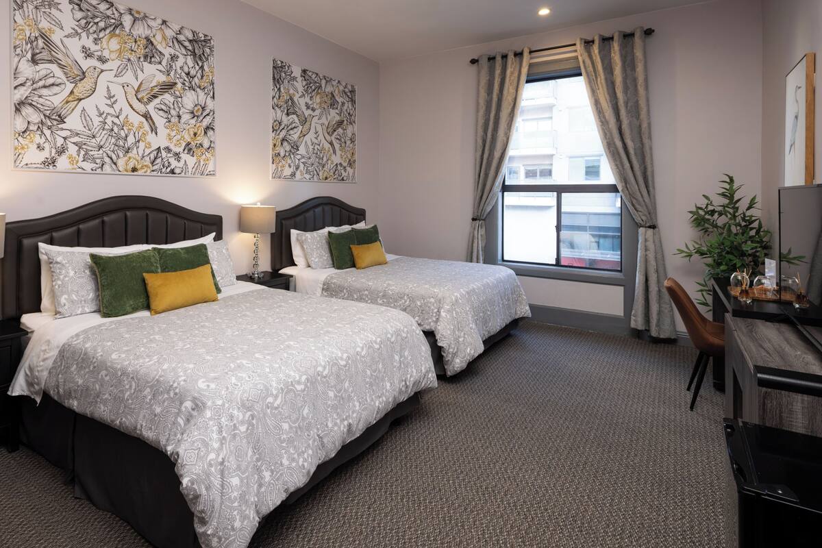 A hotel room with two beds, a desk, a chair, a window with curtains, and two large floral artwork pieces on the wall.