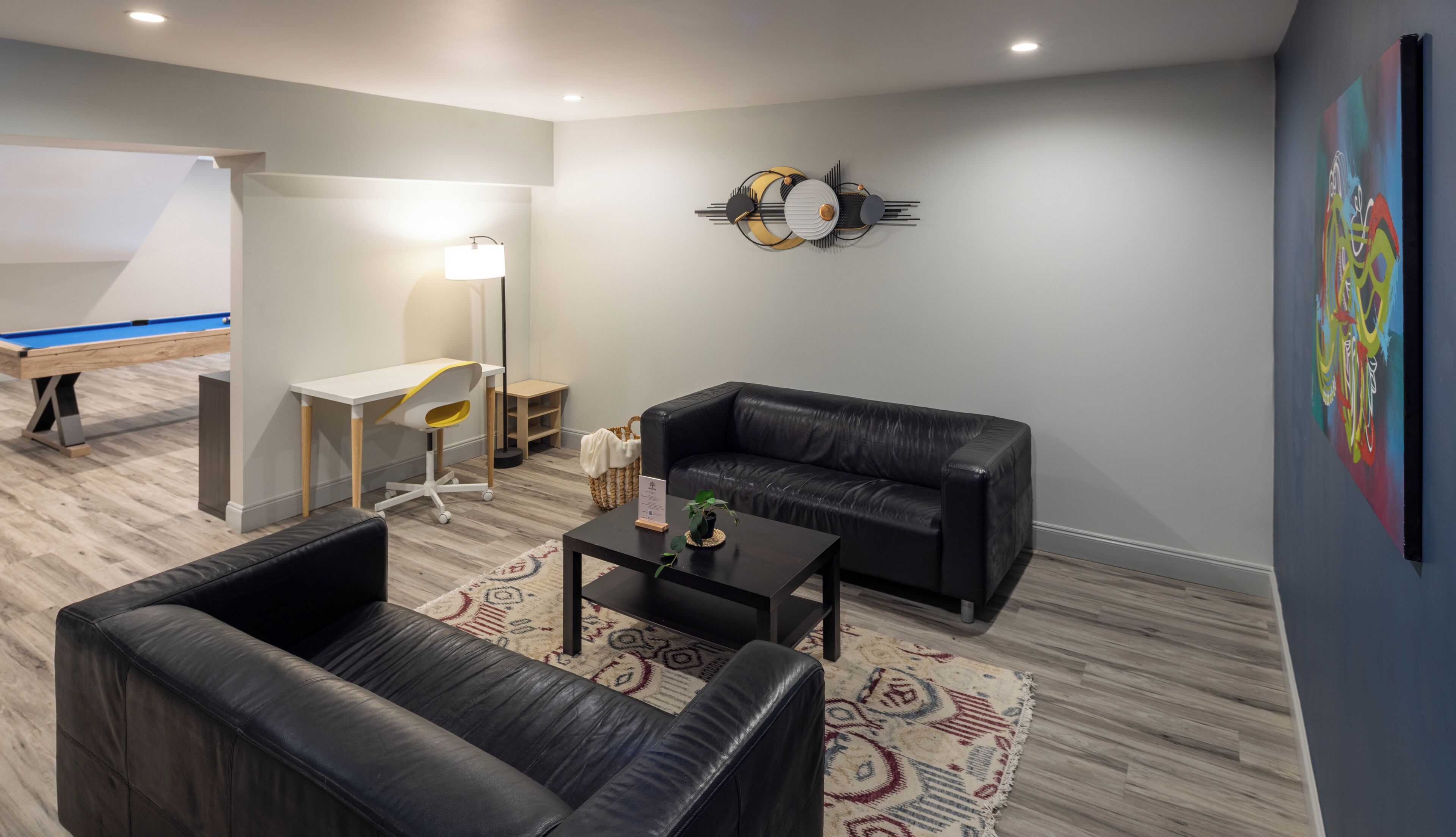A modern living room with a black leather sofa, a small coffee table, a desk with a chair, a pool table, and a wall clock.