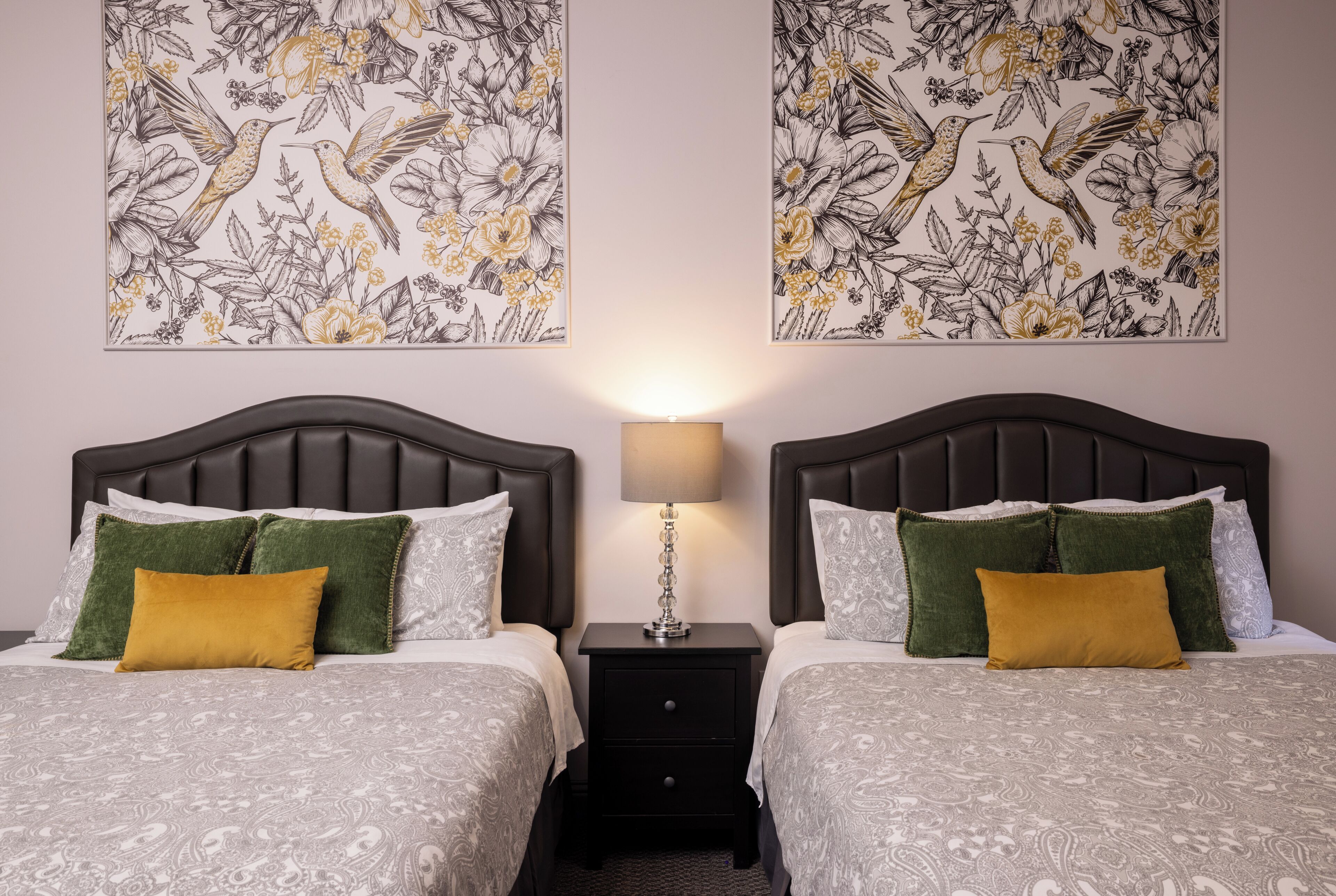 Una cama doble con una colcha estampada, dos almohadas verdes y una almohada color mostaza, una mesita de noche con una lámpara, y dos obras de arte floral colgadas en la pared.