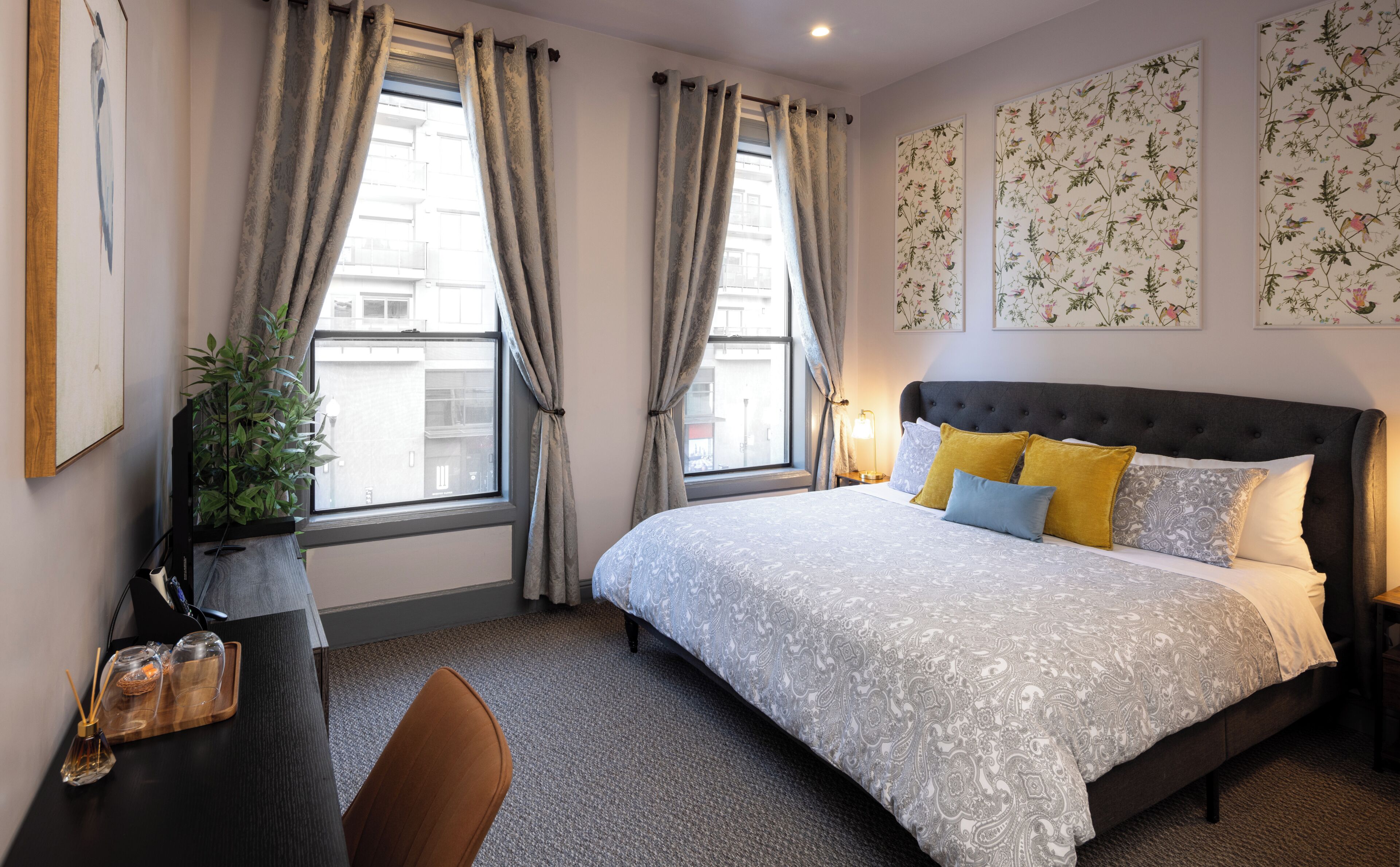 A bedroom with a bed, curtains, a plant, a desk, and wall art.