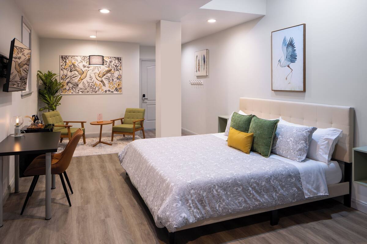 A modern hotel room with a large bed, a dining table, two chairs, a small table, and artwork on the walls.