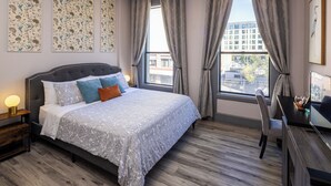 Quarto de hotel com cama, mesa de cabeceira, abajur, cadeira, escrivaninha e grandes janelas com cortinas.